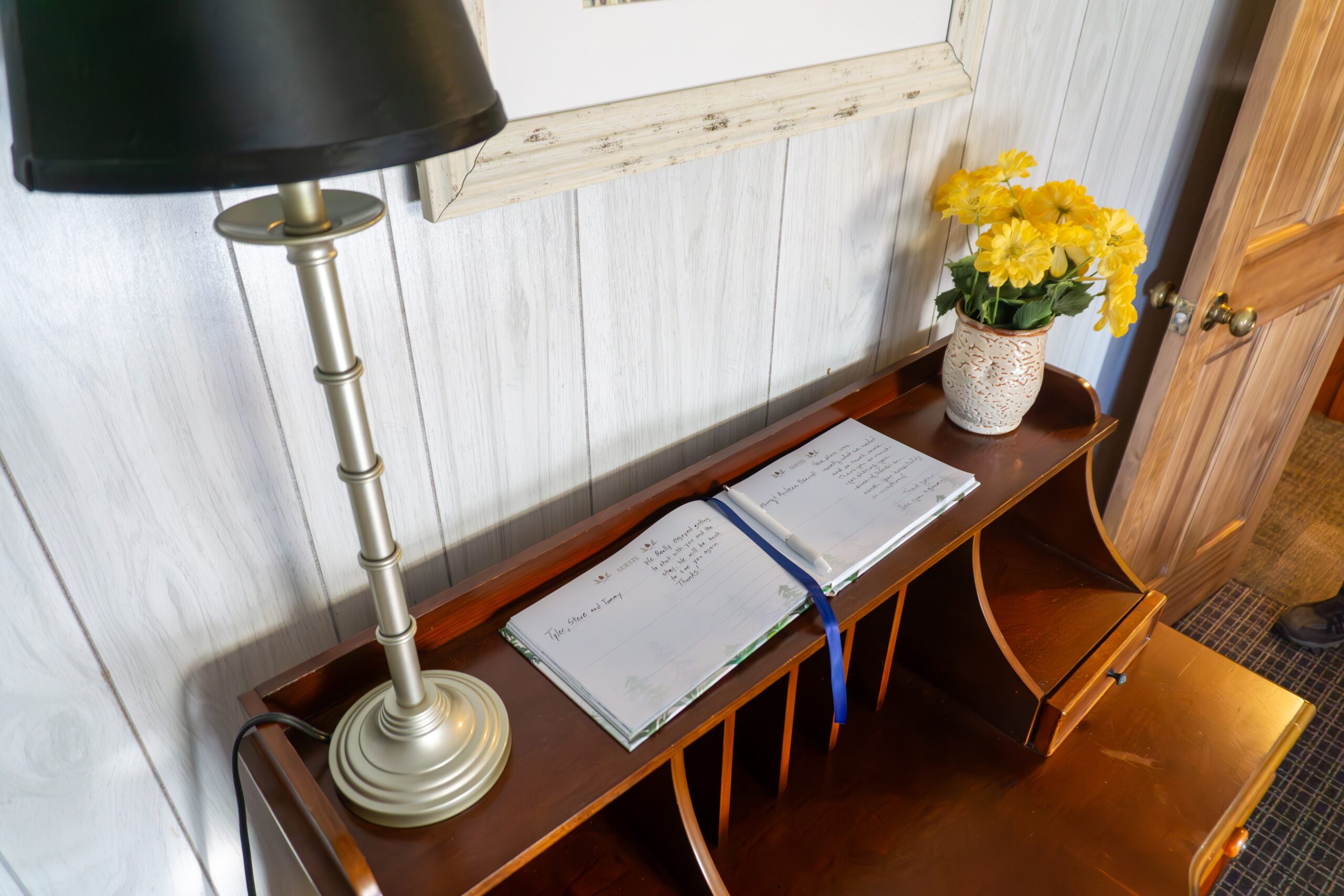 Guest book open on a wooden table at Serenity Cabins, flanked by a decorative lamp and a vase of yellow flowers, inviting guests to leave their comments.