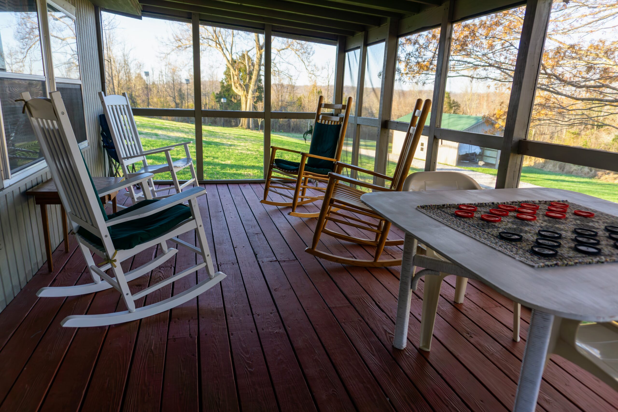 Screened front porch of Cabin #1 at Serenity Cabins, equipped with four wooden rocking chairs and a game table, overlooking a tranquil natural setting.